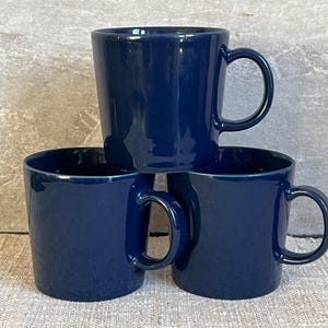 Puede incluir: Tres tazas de cerámica azul marino apiladas. Cada taza tiene un asa redondeada y una forma cilíndrica. Las tazas son de un color liso y brillante y parecen estar vacías.
