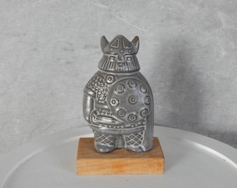 May include: A gray ceramic figurine of a Viking warrior wearing a helmet and holding a shield. The figurine is standing on a wooden base.