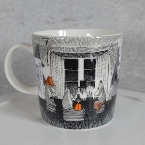 Op de afbeelding: Witte keramische mok met een zwart-witte illustratie van een scène uit de Moomin-boeken. De illustratie toont Moomin-personages die aan een tafel zitten met een raam op de achtergrond. De mok heeft een zwart handvat.