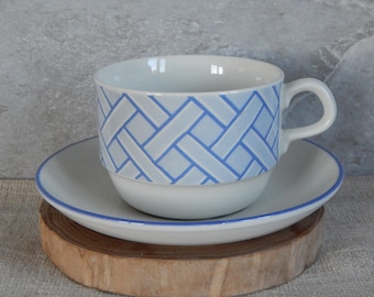 Puede incluir: Una taza de té y un platillo de cerámica blanca con un patrón tejido azul y blanco. La taza y el platillo están sobre una superficie de madera.
