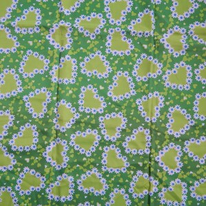 May include: Green fabric with a repeating pattern of lime green hearts outlined with white and blue daisy-like flowers. Small green and white hearts fill the background. The fabric has a bright, cheerful appearance.