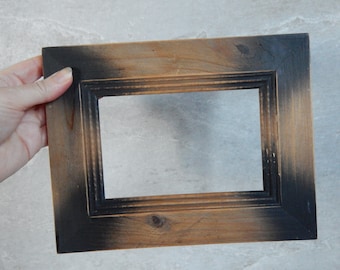 Puede incluir: Un marco de fotos rectangular de madera con un acabado envejecido. El marco tiene un borde oscuro, casi negro, que se desvanece en un marrón más claro. El marco está sostenido por una mano.