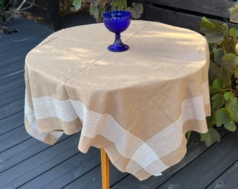 May include: A round tan tablecloth with a white striped border pattern draped over a table, featuring a single cobalt blue glass pedestal bowl with a scalloped rim placed in the center of the fabric surface.