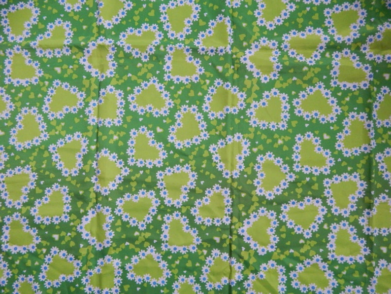 May include: Green fabric with a repeating pattern of lime green hearts outlined with white daisies and blue centers. The background is a darker green with small green hearts scattered throughout.