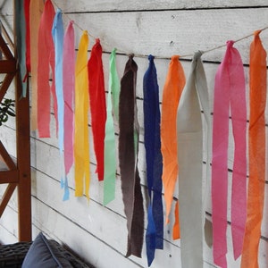 May include: A colorful fabric garland with strips of fabric in various colors, including red, orange, yellow, green, blue, and pink. The garland is hanging on a white wooden wall.