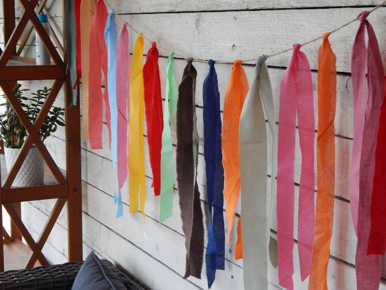 May include: A colorful fabric garland with strips of fabric in various colors, including red, orange, yellow, green, blue, and pink. The garland is hanging on a white wooden wall.