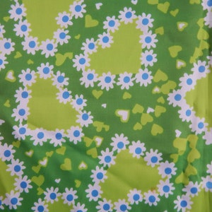 May include: A close-up of a green fabric with a repeating pattern of white daisies with blue centers, arranged in heart shapes. Small green hearts are scattered across the background. The fabric has a light green background.