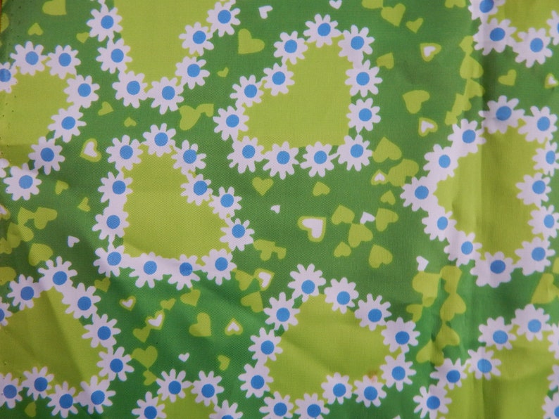 May include: A close-up of a green fabric with a repeating pattern of white daisies with blue centers, arranged in heart shapes. Small green hearts are scattered across the background. The fabric has a light green background.