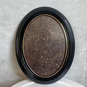 Peut inclure: Un cadre photo ovale noir avec un bord intérieur doré. Le cadre a un fond marron foncé avec des mouchetures dorées. Le cadre est posé sur une surface blanche et contre un mur gris.