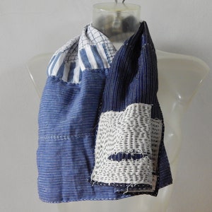 Könnte beinhalten: Ein blauer und weißer Patchwork-Schal aus recyceltem Stoff. Der Schal besteht aus verschiedenen Blautönen und Weißtönen, und die Nähte sind sichtbar. Der Schal ist in der Mitte gefaltet und wird auf einer Puppe präsentiert.