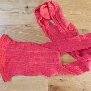 May include: A coral-colored knitted scarf with a textured pattern. The scarf is folded in half and laid flat on a wooden surface.
