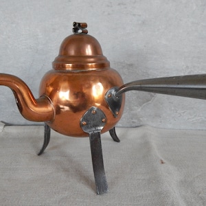 May include: A copper teapot with a domed lid and a long spout. The teapot has three iron legs and a metal handle. The teapot is on a white linen tablecloth.