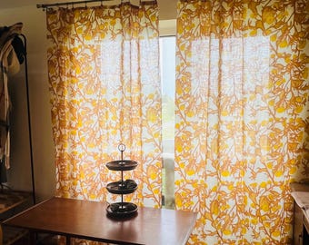 May include: A pair of floor-length curtains featuring a repeating yellow and orange botanical pattern of leaves and flowers. The curtains are hung on a thin metal rod with rings and are positioned in front of a window, allowing light to filter through.