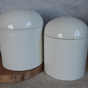 May include: Two white ceramic canisters with lids. The canisters are cylindrical and have a smooth, glossy finish. They are sitting on a wooden surface.