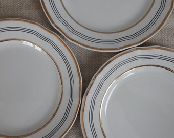 May include: Three white ceramic plates with black and gold trim. The plates have a simple design with three black lines around the edge and a thin gold line.