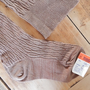May include: A pair of light brown textured socks with a ribbed design, lying on a wooden surface. The socks have a tag attached with text and measurements. The socks are made of a soft, comfortable material.