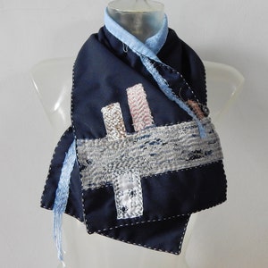 May include: A navy blue scarf with a patchwork design featuring different shades of blue, pink, and white fabric. The scarf is made of a soft, textured material and has a frayed edge.