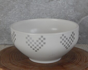 May include: White ceramic bowl with a checkered heart design in silver. The bowl is sitting on a wooden surface.