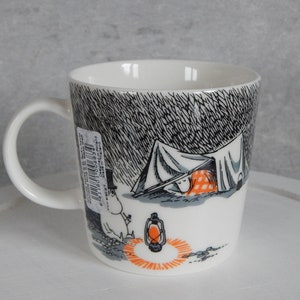 Op de afbeelding: Witte keramische mok met een zwart-witte illustratie van een personage in een tent met een lantaarn. De illustratie is omgeven door een zwart-wit patroon.