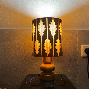 Puede incluir: Una lámpara de mesa de madera con una pantalla marrón con un patrón de hojas. La pantalla está iluminada con una luz cálida. La lámpara está sobre una superficie oscura.