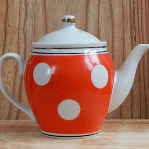 Può includere: Una teiera rossa con pois bianchi, un coperchio bianco e un bordo dorato. La teiera ha un manico e un beccuccio bianchi.
