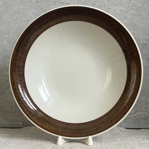 Puede incluir: Un cuenco de cerámica blanco con un borde marrón texturizado y ancho. El cuenco es redondo y se apoya en un soporte blanco. El interior del cuenco es de color blanco brillante.