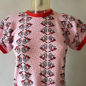 Peut inclure: Un t-shirt rose à manches courtes avec une bordure rouge autour de l'encolure et des manches. Le t-shirt présente un motif répété d'oiseaux noirs et rouges sur un fond rose avec des pois blancs.