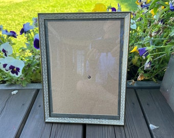 May include: A silver picture frame with a decorative border and a brown mat. The frame is sitting on a wooden surface.