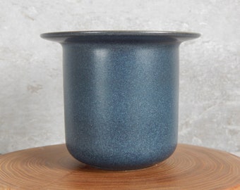 May include: A dark blue ceramic vase with a slightly flared rim. The vase has a textured surface and is sitting on a wooden surface.