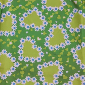 May include: A green fabric with a repeating pattern of lime green hearts outlined with white daisies and blue centers. Small green and white hearts fill the background. The fabric has a retro, floral design.