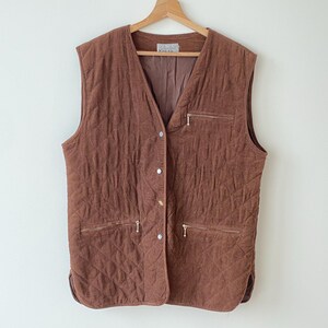 Op de afbeelding: Een bruin gewatteerd vest met een V-hals en een knoopsluiting hangt aan een houten kleerhanger. Het vest heeft twee zakken met ritssluiting en een klein label bij de kraag. De stof heeft een getextureerde uitstraling.