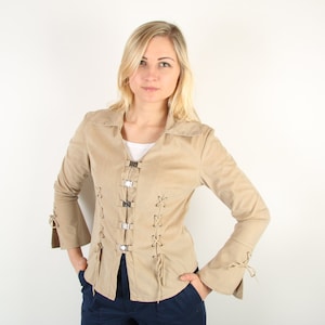 Op de afbeelding: Een beige suède jasje met een kraag, decoratieve zilveren sluitingen en uitlopende mouwen. De jas heeft vetersluiting aan de voorkant en mouwen. De persoon draagt een wit shirt en een marineblauwe broek.
