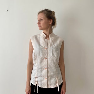 Op de afbeelding: Roze mouwloze blouse met een opstaande kraag en decoratieve knopen. De blouse heeft een subtiel bloemenpatroon en zijstrikken. Het model draagt een zwarte rok.