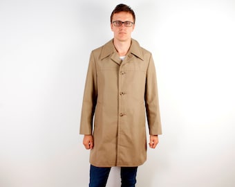 May include: A tan trench coat with a button-up front and a wide collar. The coat is knee-length and has a classic, tailored design. The coat is worn over a white shirt and blue jeans.