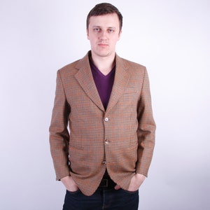 May include: A brown and orange plaid blazer with a purple v-neck shirt underneath. The blazer has two buttons and a classic notched lapel. The person is wearing dark blue jeans.