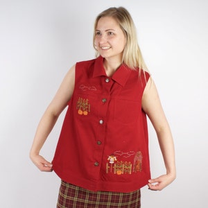 Può includere: Una camicia rossa senza maniche con bottoni e ricami a tema autunnale. Il ricamo include zucche, uno spaventapasseri e una recinzione. La camicia ha un colletto e una tasca sul petto.