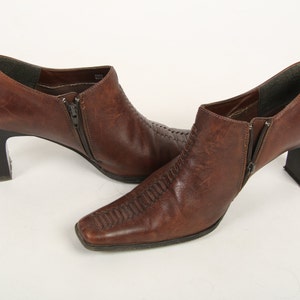 K&ouml;nnte beinhalten: Ein Paar braune Lederstiefeletten mit quadratischer Zehenpartie, Blockabsatz und seitlichem Rei&szlig;verschluss. Die Stiefel verf&uuml;gen &uuml;ber ein strukturiertes, gen&auml;htes Detail, das vertikal &uuml;ber den Fu&szlig;r&uuml;cken verl&auml;uft, mit dunkelbraunem Innenfutter.