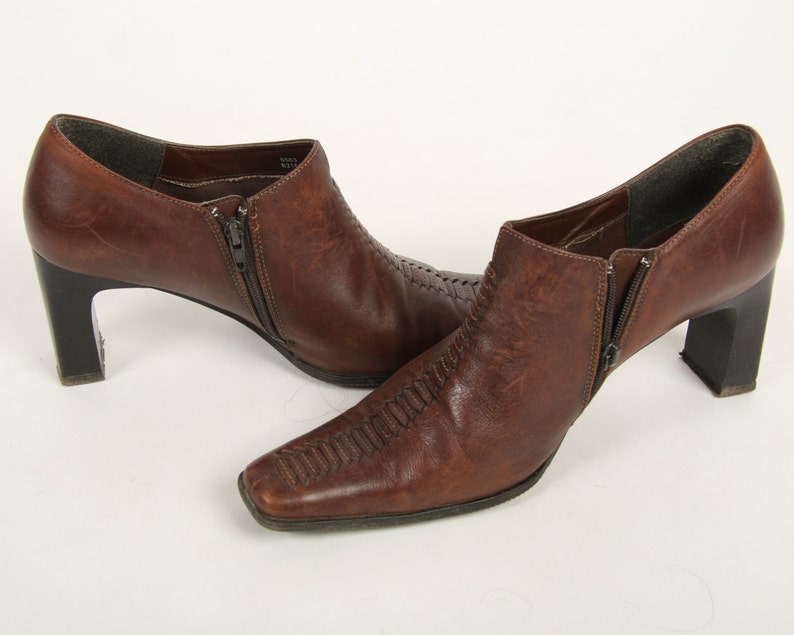 K&ouml;nnte beinhalten: Ein Paar braune Lederstiefeletten mit quadratischer Zehenpartie, Blockabsatz und seitlichem Rei&szlig;verschluss. Die Stiefel verf&uuml;gen &uuml;ber ein strukturiertes, gen&auml;htes Detail, das vertikal &uuml;ber den Fu&szlig;r&uuml;cken verl&auml;uft, mit dunkelbraunem Innenfutter.