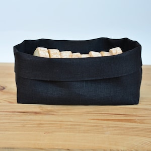 Könnte beinhalten: Ein schwarzer Brotbeutel aus Leinen, gefüllt mit mehreren Brotstücken. Der rechteckige Beutel hat einen umgeklappten oberen Rand. Der Beutel steht auf einer hellbraunen Holzoberfläche.