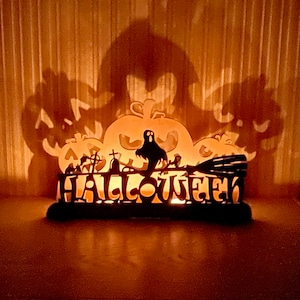 Könnte beinhalten: Ein hölzerner Halloween-Kerzenhalter mit einer Silhouette eines Geistes, Kürbissen und eines Besens. Das Wort "HALLOWEEN" ist in einer dekorativen Schrift geschrieben.