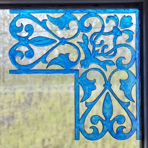 Pode incluir: Um canto de uma janela ou painel decorativo com um design azul brilhante e ornamentado. O padrão inclui elementos florais e de rolagem estilizados, emoldurados por uma borda preta. O fundo mostra uma vista desfocada da folhagem externa.