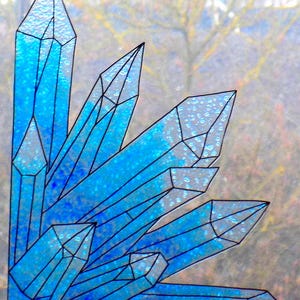 May include: A stained glass window panel depicting a cluster of blue crystals with black outlines. The crystals have a gradient of light to dark blue, with a textured, sparkling appearance. The background is a blurred view of a tree.
