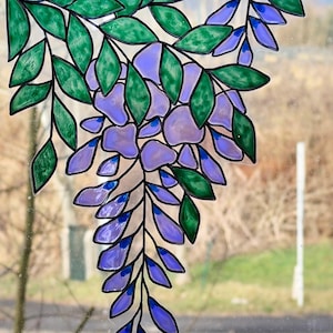 May include: Stained glass window art featuring a wisteria design. The artwork showcases green leaves and purple and blue flower clusters, outlined in black. The window art is a decorative piece for home decor.