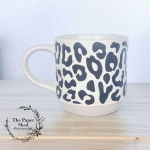 May include: A white ceramic mug with a black leopard print design. The mug has a rounded handle and a wide base.