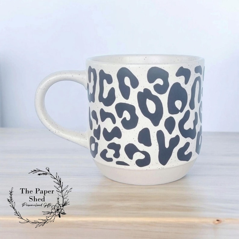 May include: A white ceramic mug with a black leopard print design. The mug has a rounded handle and a wide base.