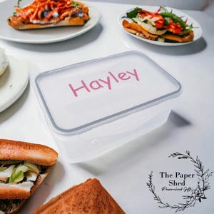 May include: A clear plastic rectangular food container with a pink label that says "Hayley". The container is on a white surface. The Paper Shed Personalised Gifts is printed in black on a white background with a floral design.