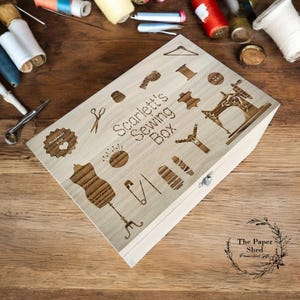 May include: A wooden box with a hinged lid, engraved with the words "Scarlett's Sewing Box" and various sewing-related icons. The box is light brown and has a silver latch. The text "Handmade with love" is also engraved on the box.
