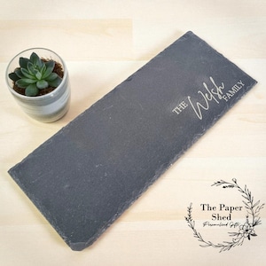 May include: A dark grey slate serving board, rectangular in shape, engraved with the words "The Welsh Family". A small succulent plant in a striped ceramic pot is visible. The Paper Shed logo is in the lower right corner.