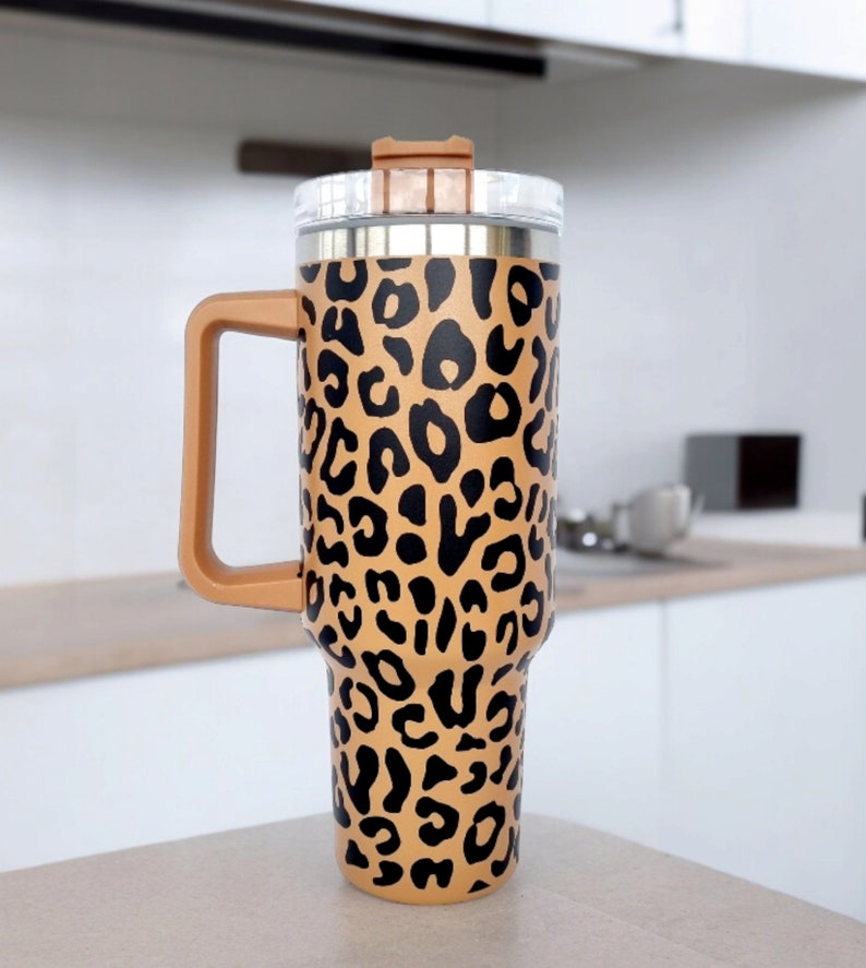 May include: A stainless steel travel mug with a brown and black leopard print design. The mug has a lid and a handle.