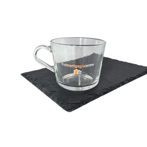 May include: A clear glass teacup with a handle, featuring the text "themortgagecentre" in black. The cup sits on a dark grey rectangular slate coaster. The logo includes orange and yellow elements.